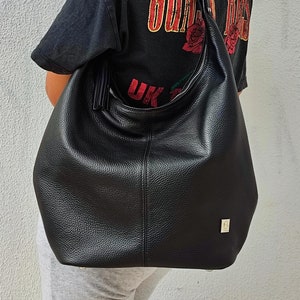 May include: A large, black leather shoulder bag with a curved zipper and a long strap. The bag has a textured surface and a small gold-colored square emblem. The person in the image is wearing a black t-shirt with the text "GUNS N' ROSES" and a red rose graphic.