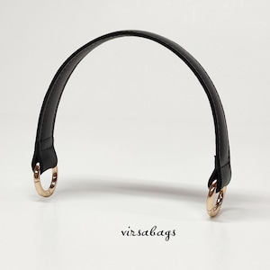 May include: A black arched handbag strap featuring stitched edges and polished gold-tone circular metal clasps at both ends. The text virsabags is printed in a small black cursive font below the center of the arched strap.