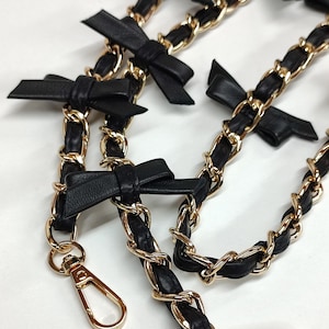 May include: A black leather chain strap with gold chain links and black leather bows. The strap has two gold clasps.