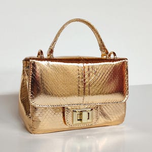 Puede incluir: Un pequeño bolso de mano dorado con un patrón texturizado similar a la piel de serpiente. El bolso tiene un asa superior, una forma rectangular y un cierre de broche. Los herrajes también son dorados.