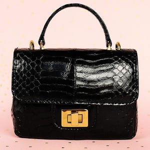 Op de afbeelding: Een kleine, zwarte handtas met een textuur, krokodilachtig patroon. De tas heeft een handvat aan de bovenkant, goudkleurige hardware en een rechthoekige sluiting. De achtergrond is lichtroze met gouden confetti.