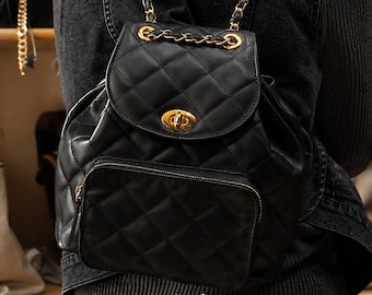 May include: Black quilted leather backpack with gold-tone chain straps and hardware. The backpack features a flap closure with a turn-lock clasp and a front zippered pocket. The bag is worn on the back of a person wearing a black denim jacket.