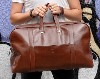 Puede incluir: Un bolso de viaje rectangular grande de cuero marrón con dos asas superiores, acabado liso y detalles de costuras verticales. El bolso cuenta con un cierre de cremallera central, detalles de herrajes metálicos y un pequeño logotipo grabado.