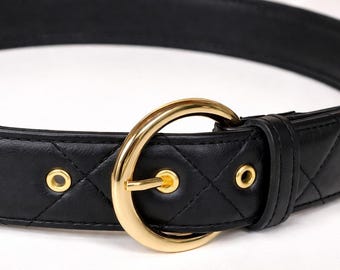 May include: A black quilted leather belt with a gold-tone buckle. The belt features a diamond pattern and gold rivets. The buckle is a large, circular design. This belt is a fashion accessory.