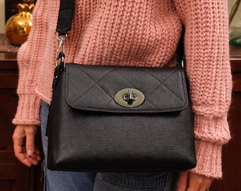 May include: Black leather crossbody bag with a quilted flap and silver-tone clasp. The bag has a black adjustable strap. The bag is worn over a pink knit sweater and blue jeans.