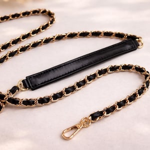 May include: A replacement handbag strap featuring a gold-tone metal chain interwoven with a black leather-like strip. The strap includes a flat, solid black shoulder pad section and a gold-tone metal lobster clasp at the end for secure attachment.