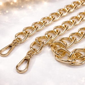 Peut inclure: Gros plan de plusieurs chaînes dorées avec des fermoirs mousquetons. Les chaînes sont composées de maillons ovales imbriqués. Le fond est un blanc doux et flou avec des reflets subtils, créant une sensation de luxe. Idéal pour les sacs à main.