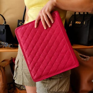 May include: A rectangular, hot pink clutch bag featuring a textured, pebbled surface with diagonal quilted stitching. The bag has a slim, flat profile and a zippered closure along the top edge, showcasing a vibrant, solid color throughout the exterior.