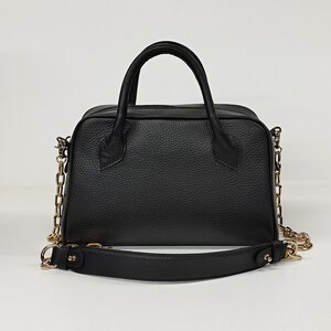 May include: A black leather handbag with a structured shape. It features a top handle and a detachable shoulder strap. The bag has gold-colored chain accents and hardware. The leather has a textured grain.