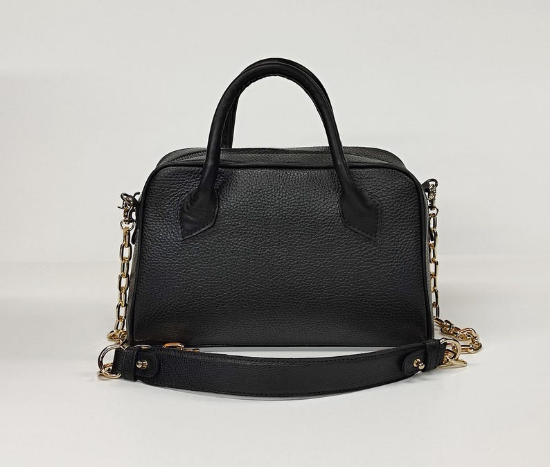 May include: A black leather handbag with a structured shape. It features a top handle and a detachable shoulder strap. The bag has gold-colored chain accents and hardware. The leather has a textured grain.