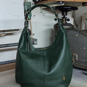 May include: A dark green hobo bag with a pebbled texture, a single shoulder strap attached with gold-tone hardware, a matching leather tassel, and a small gold-tone rectangular logo plaque on the lower front corner. The bag features a central seam.