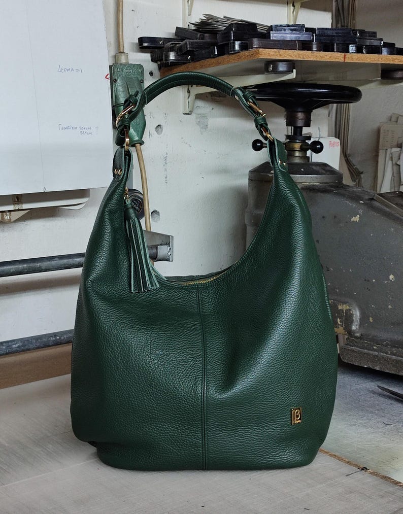May include: A dark green hobo bag with a pebbled texture, a single shoulder strap attached with gold-tone hardware, a matching leather tassel, and a small gold-tone rectangular logo plaque on the lower front corner. The bag features a central seam.