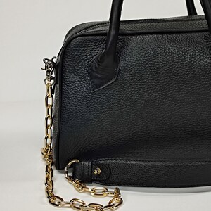 May include: Black leather handbag with a gold chain strap and handles. The bag has a textured surface and a zipper closure. The gold chain is attached to the bag with metal clasps. The bag is a medium size.