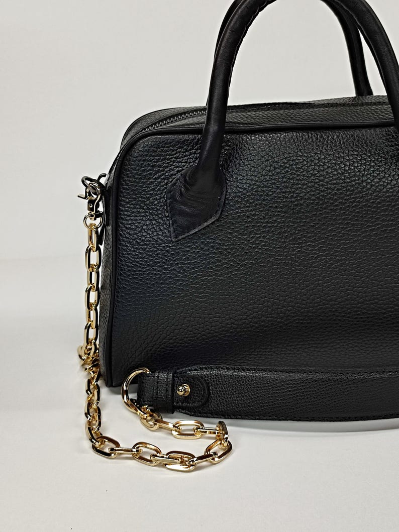 May include: Black leather handbag with a gold chain strap and handles. The bag has a textured surface and a zipper closure. The gold chain is attached to the bag with metal clasps. The bag is a medium size.
