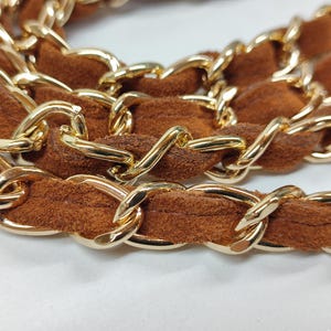 May include: A handbag strap featuring a series of interlocking gold-tone metal chain links woven with a soft, textured brown suede material. The strap is arranged in a coiled, layered formation, highlighting the contrast between the metallic finish.