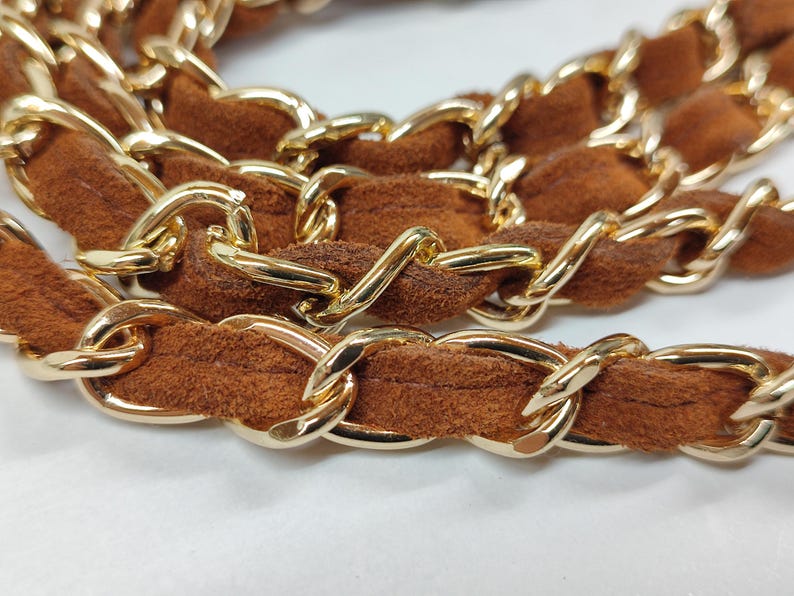 May include: A handbag strap featuring a series of interlocking gold-tone metal chain links woven with a soft, textured brown suede material. The strap is arranged in a coiled, layered formation, highlighting the contrast between the metallic finish.