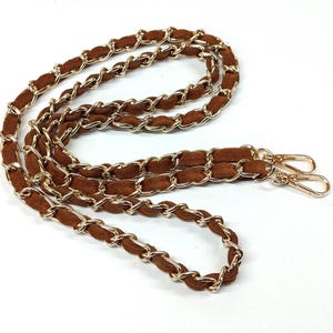 May include: A brown suede strap interwoven with gold-tone metal chain links. The strap features two gold-tone lobster claw clasps at the ends, designed for attaching to a bag or accessory. The chain and suede are linked in a continuous loop.