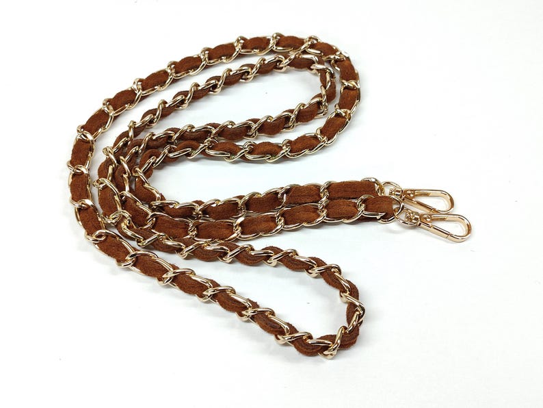May include: A brown suede strap interwoven with gold-tone metal chain links. The strap features two gold-tone lobster claw clasps at the ends, designed for attaching to a bag or accessory. The chain and suede are linked in a continuous loop.