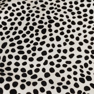 May include: A textile surface featuring a high-contrast pattern of irregular, solid black spots distributed across a creamy off-white background. The texture appears short and dense, creating a repeating spotted motif across the entire visible area.