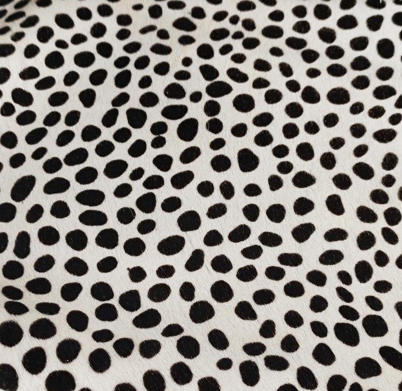 May include: A textile surface featuring a high-contrast pattern of irregular, solid black spots distributed across a creamy off-white background. The texture appears short and dense, creating a repeating spotted motif across the entire visible area.