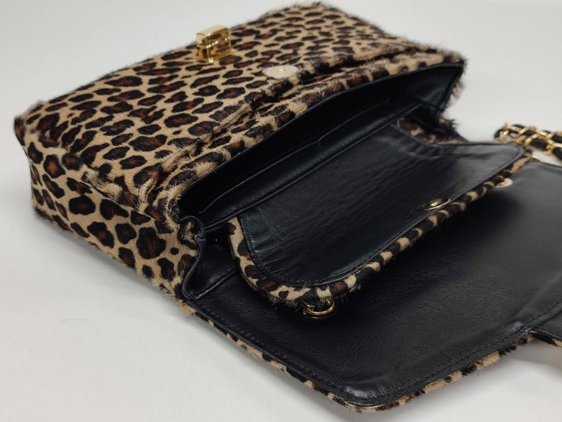 May include: A rectangular shoulder bag featuring a textured leopard print pattern in shades of brown and black. The bag has a gold-toned metal clasp closure on the front flap and a black interior lining with a gold chain strap attachment.