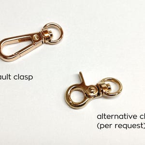 May include: Two gold-toned metal clasps for bags or purses. The clasp on the left is a traditional hook and loop style. The clasp on the right is a more modern, circular style. The text "default clasp" is below the left clasp and the text "alternative clasp (per request)" is below the right clasp.