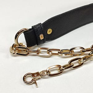 May include: Close-up of a black leather strap with gold-colored metal hardware. The strap has a textured surface and is connected to a gold chain. A gold-colored clasp is visible at the end of the chain. The hardware includes a D-ring and rectangular accents.