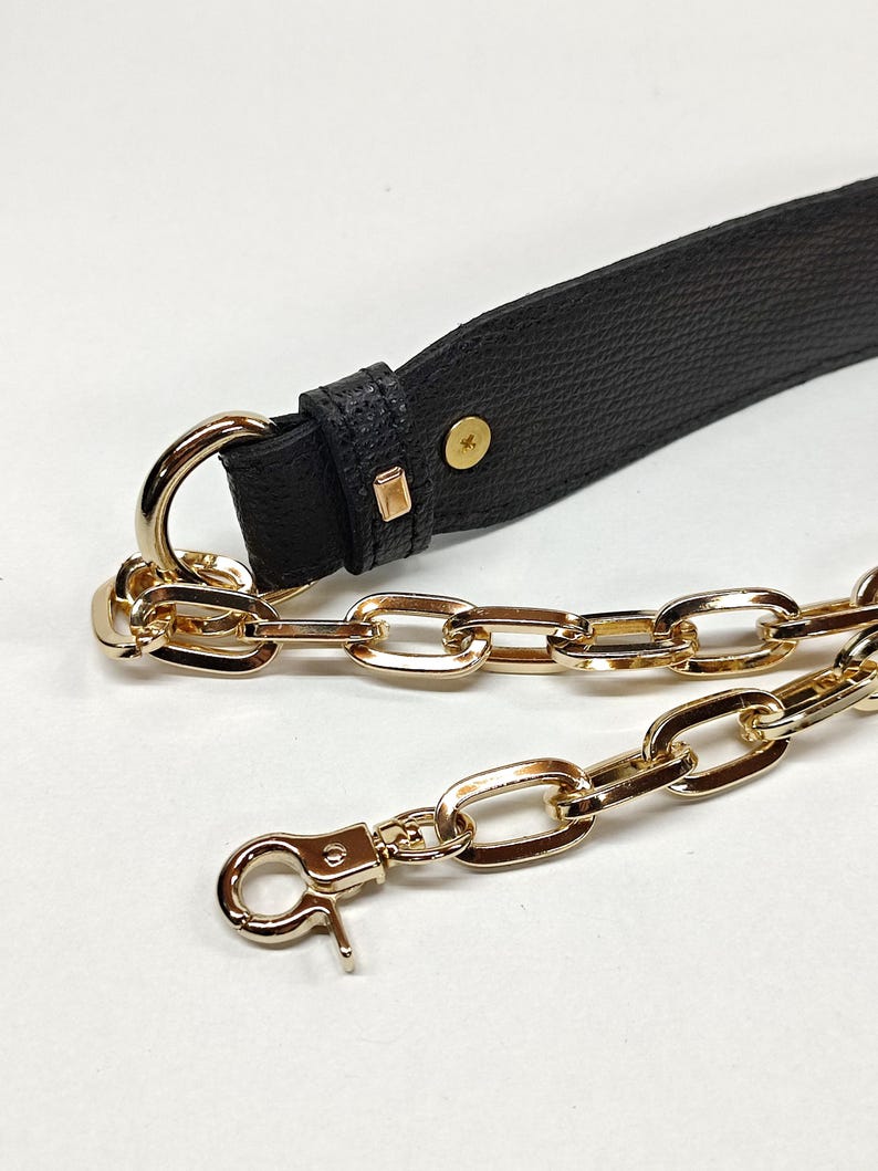 May include: Close-up of a black leather strap with gold-colored metal hardware. The strap has a textured surface and is connected to a gold chain. A gold-colored clasp is visible at the end of the chain. The hardware includes a D-ring and rectangular accents.
