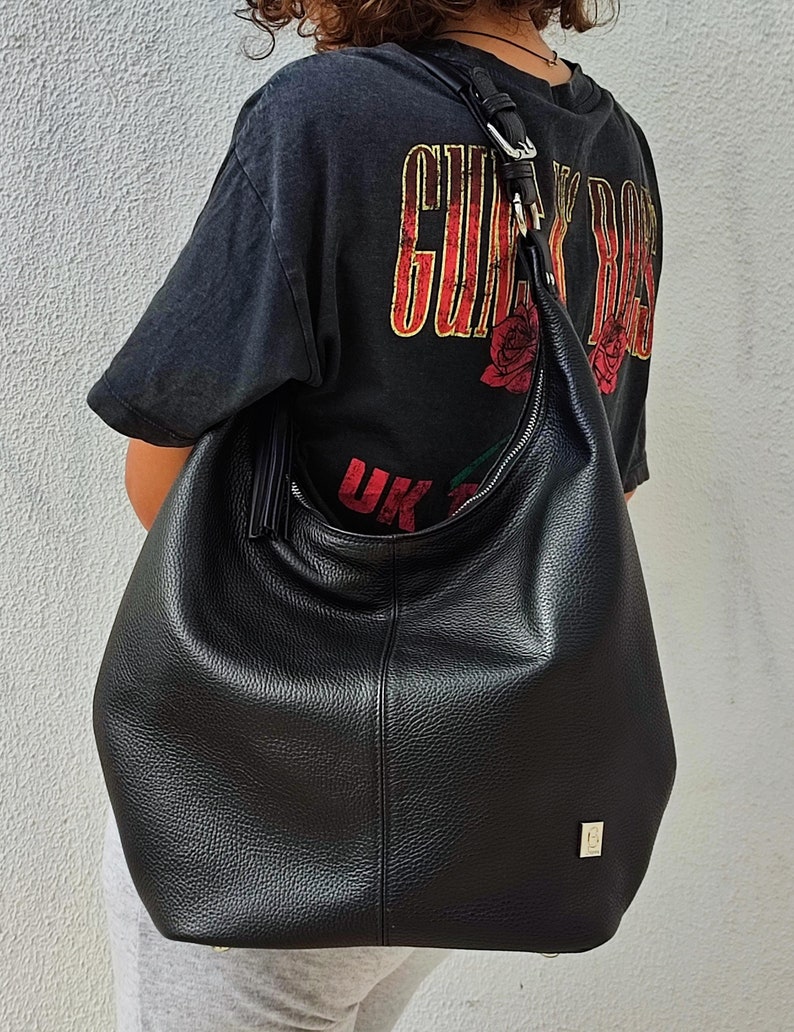 May include: A large, black leather shoulder bag with a curved zipper and a long strap. The bag has a textured surface and a small gold-colored square emblem. The person in the image is wearing a black t-shirt with the text "GUNS N' ROSES" and a red rose graphic.