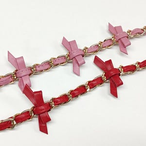 K&ouml;nnte beinhalten: Zwei Kettenarmb&auml;nder mit goldfarbenen Gliedern. Eines ist rosa mit rosa Schleifen, das andere rot mit roten Schleifen. Die Schleifen sind gleichm&auml;&szlig;ig &uuml;ber die Armb&auml;nder verteilt.