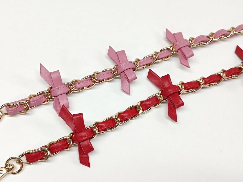 K&ouml;nnte beinhalten: Zwei Kettenarmb&auml;nder mit goldfarbenen Gliedern. Eines ist rosa mit rosa Schleifen, das andere rot mit roten Schleifen. Die Schleifen sind gleichm&auml;&szlig;ig &uuml;ber die Armb&auml;nder verteilt.