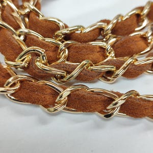 May include: A handbag strap featuring a polished gold-tone metal chain interwoven with a soft, brown suede-like material. The chain links are oval-shaped and connected in a continuous, repeating pattern throughout the length of the strap.