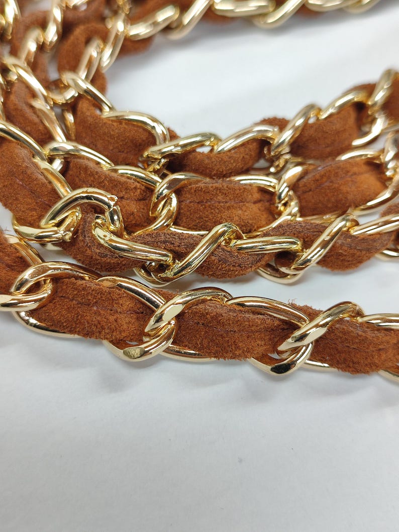 May include: A handbag strap featuring a polished gold-tone metal chain interwoven with a soft, brown suede-like material. The chain links are oval-shaped and connected in a continuous, repeating pattern throughout the length of the strap.