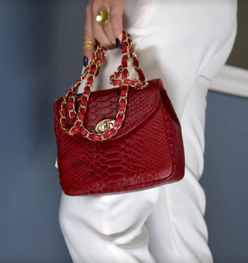 May include: A red handbag with a snakeskin texture. The bag has a gold chain strap with red leather accents. The clasp is gold-colored. The bag is held by a person wearing gold rings and a large gold ring.