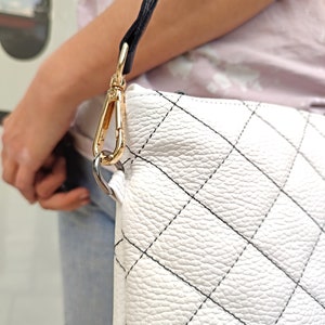 May include: A white quilted handbag featuring a textured leather-like surface with a diamond-shaped stitched pattern in black thread. The bag includes a gold-toned metal lobster clasp hardware that connects a black strap to the white bag body.