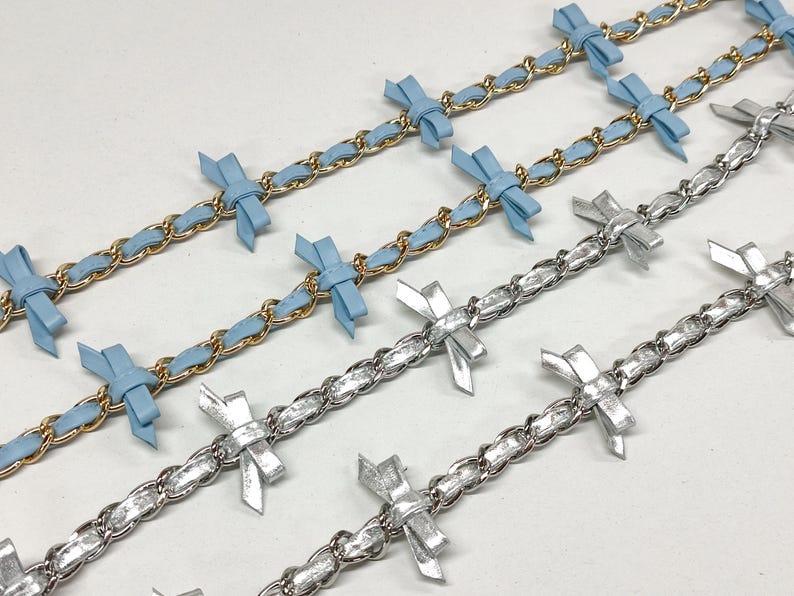 K&ouml;nnte beinhalten: Nahaufnahme mehrerer dekorativer Ketten. Zwei Ketten haben ein goldfarbenes Kettenglied-Design mit hellblauen B&auml;ndern und Schleifen. Die anderen Ketten haben ein silberfarbenes Kettenglied-Design mit silbernen Schleifen.