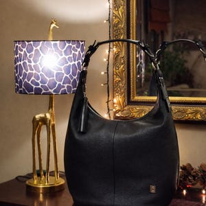 May include: A black hobo-style shoulder bag crafted from textured leather. The bag features a single curved handle attached with silver-toned hardware, a decorative leather tassel pull on the side zipper, and a small metallic logo plaque on the front.