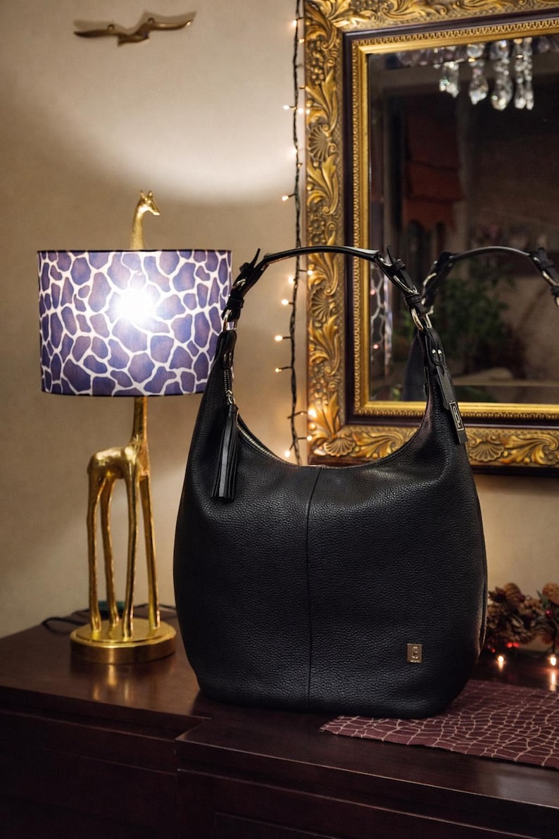 May include: A black hobo-style shoulder bag crafted from textured leather. The bag features a single curved handle attached with silver-toned hardware, a decorative leather tassel pull on the side zipper, and a small metallic logo plaque on the front.