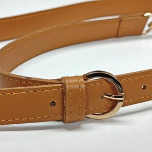 May include: A tan leather strap featuring a polished gold-tone circular buckle and matching gold-tone hardware. The strap includes visible stitching along the edges and a leather loop to secure the end of the strap through the buckle.