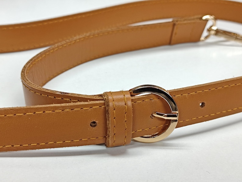 May include: A tan leather strap featuring a polished gold-tone circular buckle and matching gold-tone hardware. The strap includes visible stitching along the edges and a leather loop to secure the end of the strap through the buckle.
