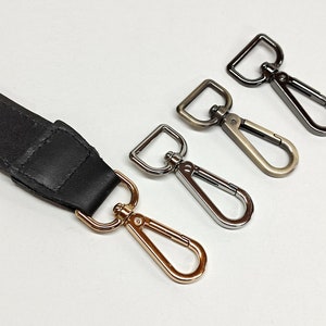 May include: A black leather strap attached to a gold-toned swivel lobster clasp, accompanied by three additional detached swivel lobster clasps in silver, antique brass, and gunmetal finishes, each featuring a rectangular loop attachment.