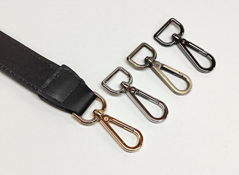 May include: A black leather strap attached to a gold-toned swivel lobster clasp, accompanied by three additional detached swivel lobster clasps in silver, antique brass, and gunmetal finishes, each featuring a rectangular loop attachment.