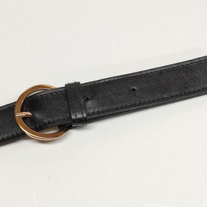 May include: A black belt featuring a smooth texture and a polished gold-tone circular buckle. The belt includes a matching black leather-like keeper loop and pre-punched holes along the strap for adjustable fastening and secure closure.