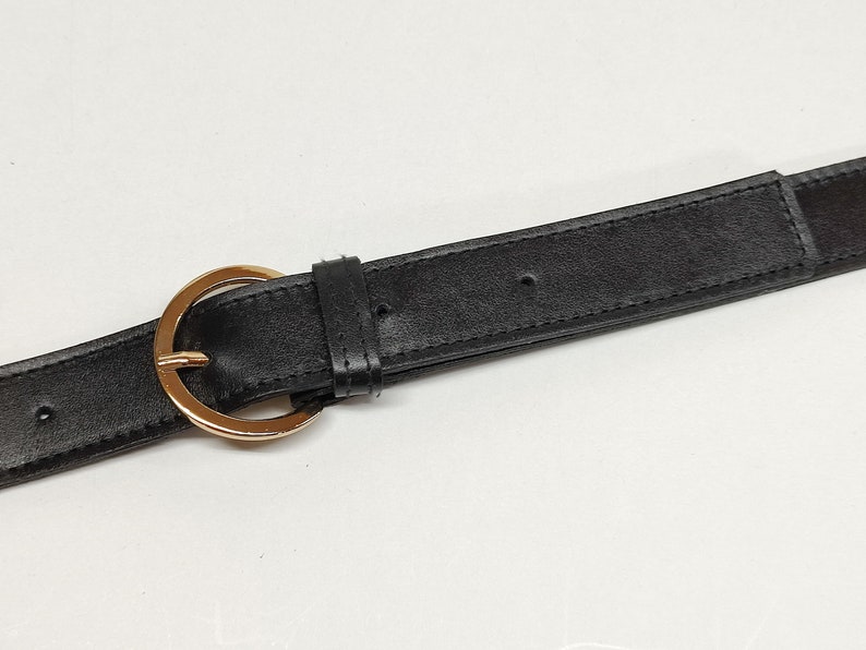 May include: A black belt featuring a smooth texture and a polished gold-tone circular buckle. The belt includes a matching black leather-like keeper loop and pre-punched holes along the strap for adjustable fastening and secure closure.