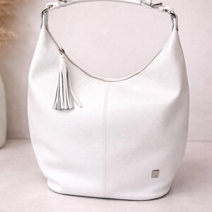 May include: A white hobo bag with a textured surface, featuring a single shoulder strap attached with silver-toned hardware, a top zipper closure with a matching leather tassel pull, and a small square silver-toned logo plaque on the lower front.