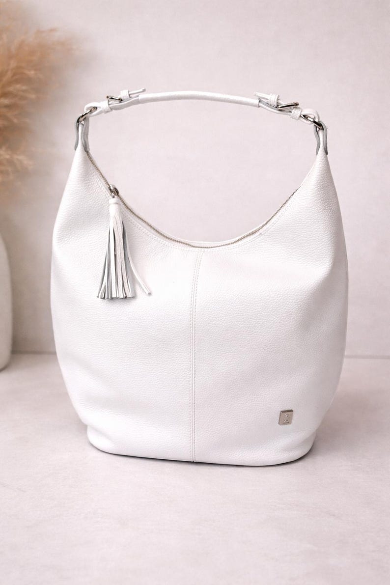 May include: A white hobo bag with a textured surface, featuring a single shoulder strap attached with silver-toned hardware, a top zipper closure with a matching leather tassel pull, and a small square silver-toned logo plaque on the lower front.