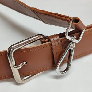 May include: A brown leather strap featuring a silver-toned metal rectangular buckle and a matching silver-toned lobster clasp. The strap is constructed with visible stitching along the edges, showcasing the smooth texture of the leather material.