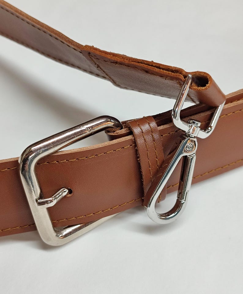 May include: A brown leather strap featuring a silver-toned metal rectangular buckle and a matching silver-toned lobster clasp. The strap is constructed with visible stitching along the edges, showcasing the smooth texture of the leather material.