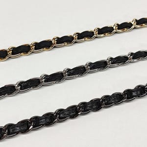 May include: Three metal chain straps for bags, each featuring a black leather or faux leather strip woven through the links. The top chain is gold-toned, the middle chain is silver-toned, and the bottom chain is a dark gunmetal-toned finish.