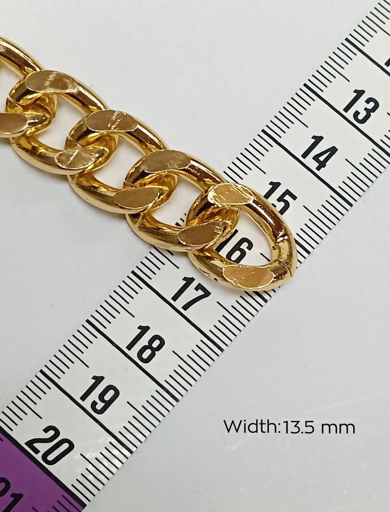 Puede incluir: Eslab&oacute;n de cadena de oro con una superficie plana ligeramente texturizada. La cadena se mide con una regla, mostrando un ancho de 13,5 mm.
